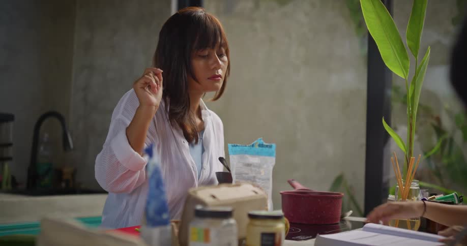 Two women are in a kitchen at midday one dressed in white is whisking an egg on a plate held in her arm while checking food in a pot on the stove as the other woman looks through a book