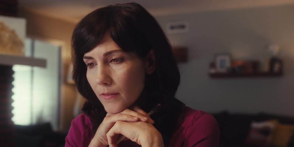 A woman sits indoors with her hands under her chin gazing thoughtfully off camera in a softly lit room