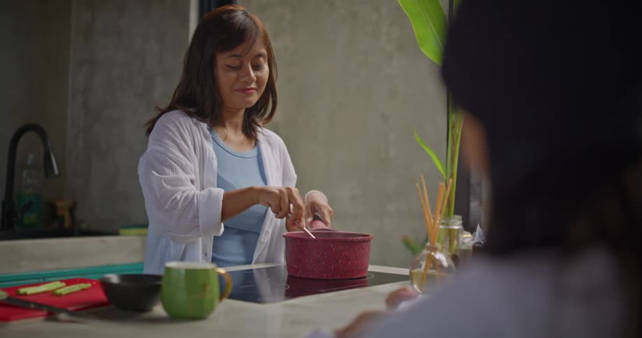 A woman in a kitchen smiling and stirring a pot with a spoon while talking to other women who are watching her