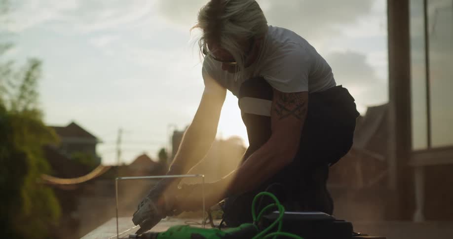 A person with short blonde hair and tattoos on their left arm uses a power tool to work on a surface outdoors