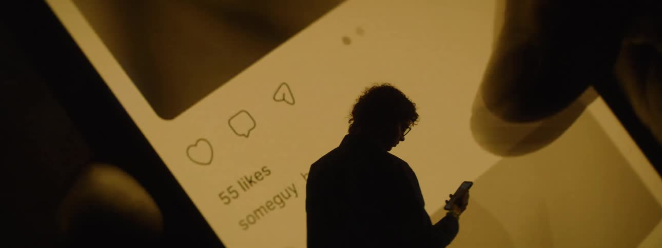 A man standing and looking at his phone screen while in front of him theres a large screen displaying a social media app interface mirroring a smartphone