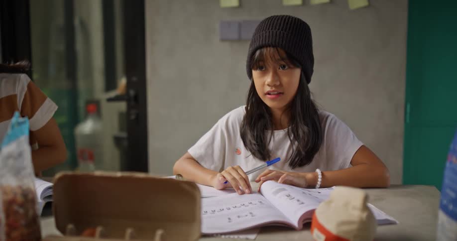 A child at school holding a pen reading a book and talking to a teacher next to the child a teenager is also studying