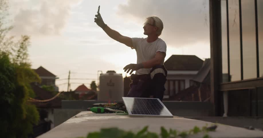 A man on a rooftop holds up his phone while checking for signal with a small solar panel and tools beside him at sunset