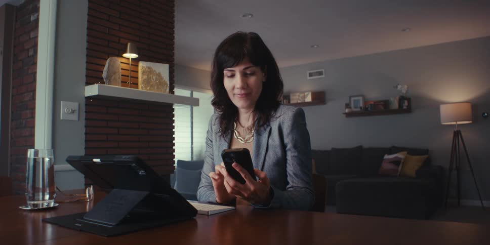 A woman in a business suit sits at a table focused on her phone in a modern living room