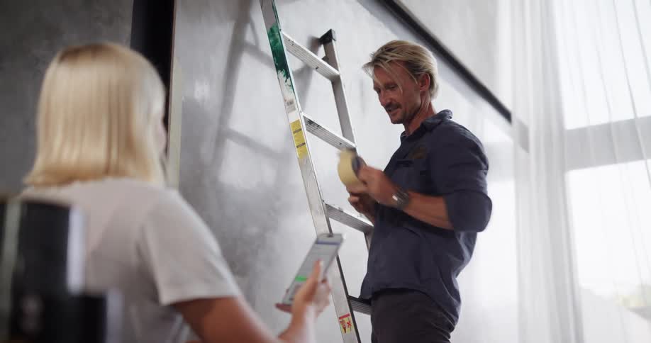Man on a ladder carrying a masking tape and talking to a woman next to him