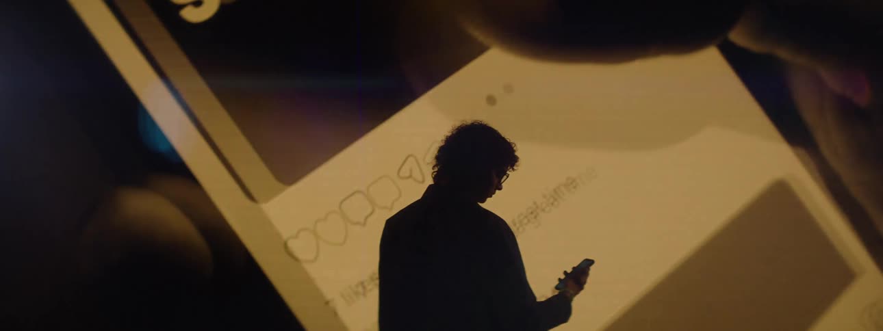 Silhouette of a man interacting with a smartphone facing a large screen that displays hands interacting with the cell phone