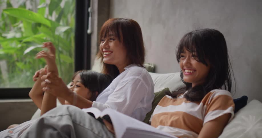 Three asian girls sit together on a cozy couch near a window smiling and laughing while spending relaxed time together