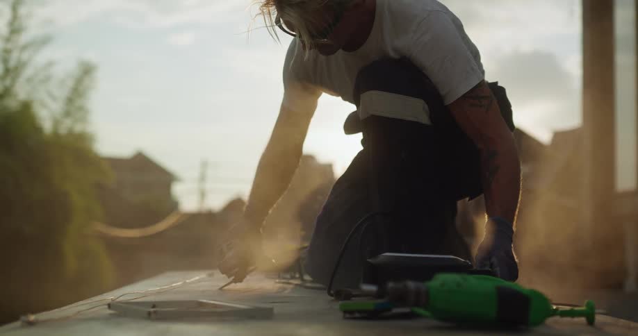 A man kneels on a deck he is building while grooving with a power tool