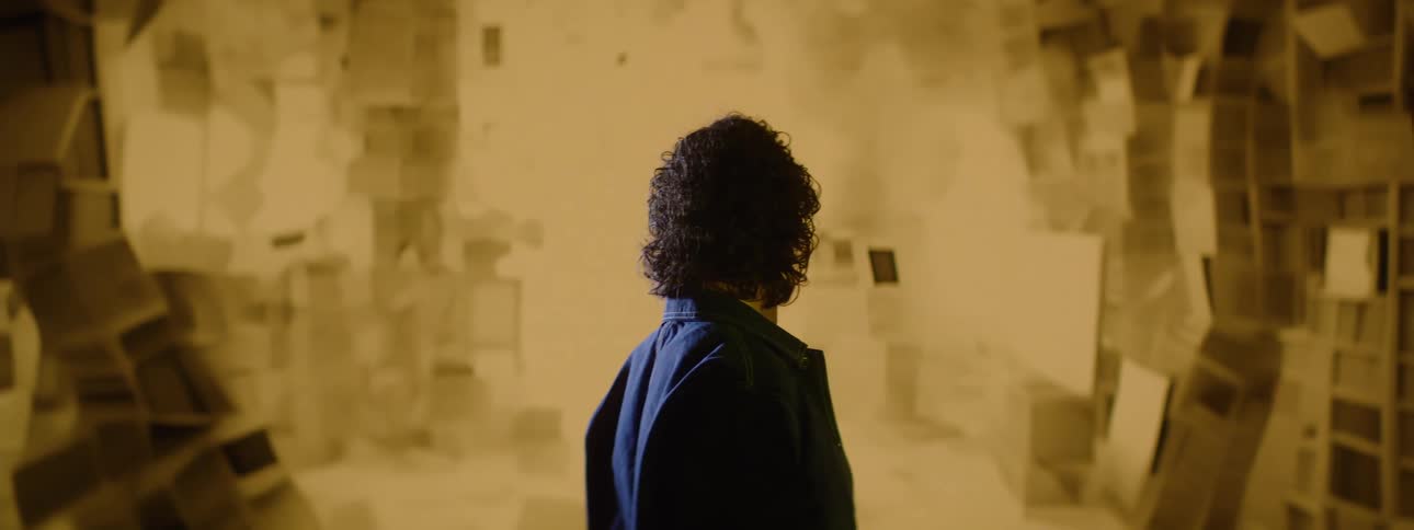 A person with dark curly hair in a dark jacket stands looking into a hazy sepia toned environment filled with abstract out of focus shapes