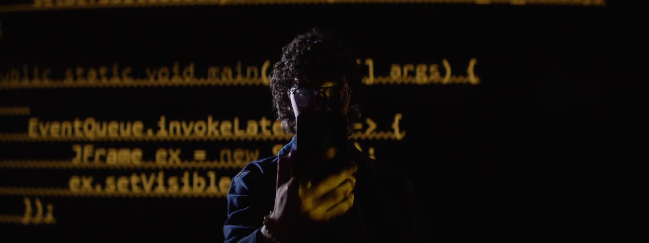 A man looks at his cellphone as a screen behind him flashes a lot of text