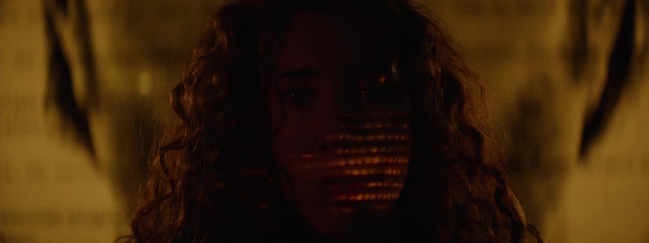 In a shadowy warm toned close up a curly haired woman turns from profile to look directly at the camera as shifting patterns of light and shadow play across her face