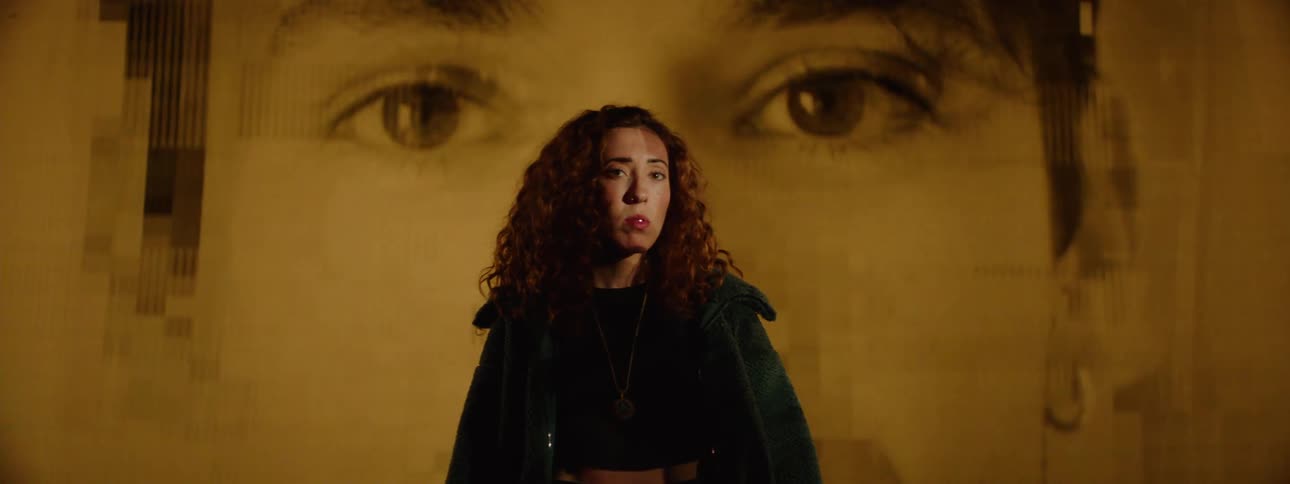 A woman stands pensively in front of a large projected image of a persons eyes as the frames behind her subtly change