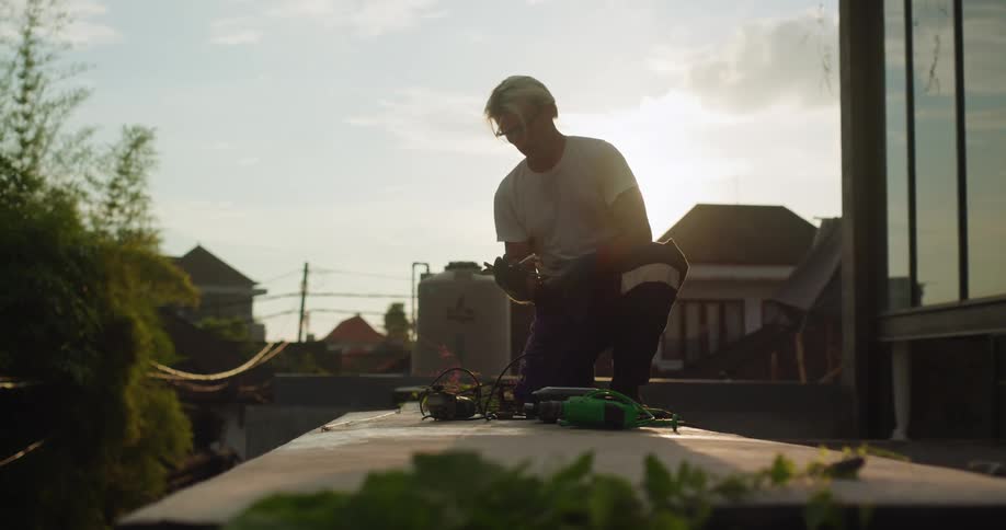 A person works on a rooftop at sunset bending over tools and cables with buildings silhouetted in the background