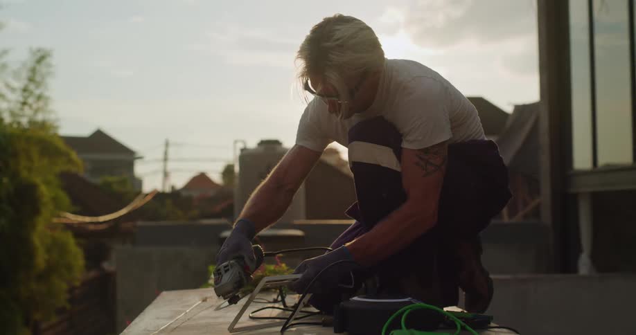 A man is kneeling on a rooftop while using a power tool to install or repair a metal structure under the setting sun