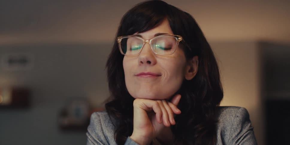 A woman in glasses thoughtfully rests her chin on her hand while looking offscreen in a softly lit indoor setting