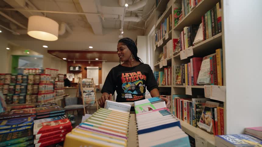 A woman in a bookstore pushing a cart with books and looking at the other books on the shelf