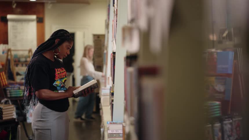 A woman is placing or retrieving a book from a tall bookshelf in a bookstore or library setting