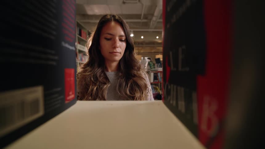 A woman browses through a library or bookstore viewed from between two large books on a shelf