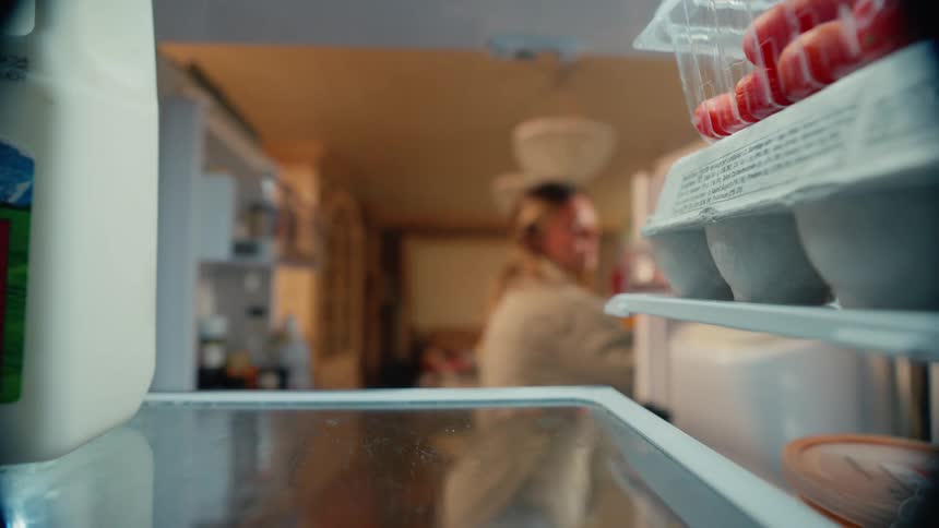 A woman reaches into a refrigerator and grabs bell peppers seen from inside the fridge