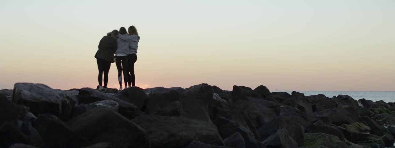 Three women standing on a rocky cliff looking at the sunset