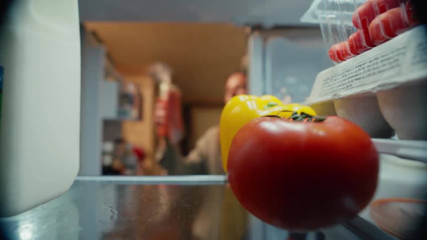 A woman opens a refrigerator reaches in and grabs a bag of carrots before closing the door