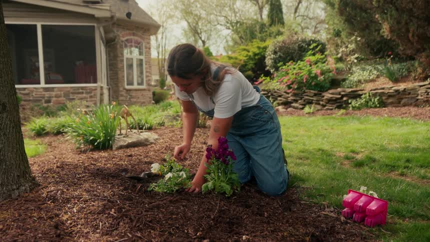 A woman is kneeling in a garden bed planting flowers in the yard of a suburban home on a sunny day