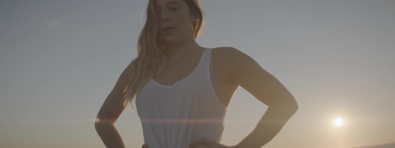 A woman is standing with hands on hips and breathing deeply as the sun sets