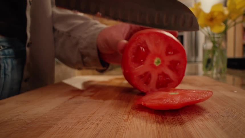 Close up of big, red, juicy tomatoes being placed on a cutting board on a kitchen counter top with a vase of fresh flowers, and then the hands a woman slicing the tomato with a large Santoku kitchen knife as the camera pans back.