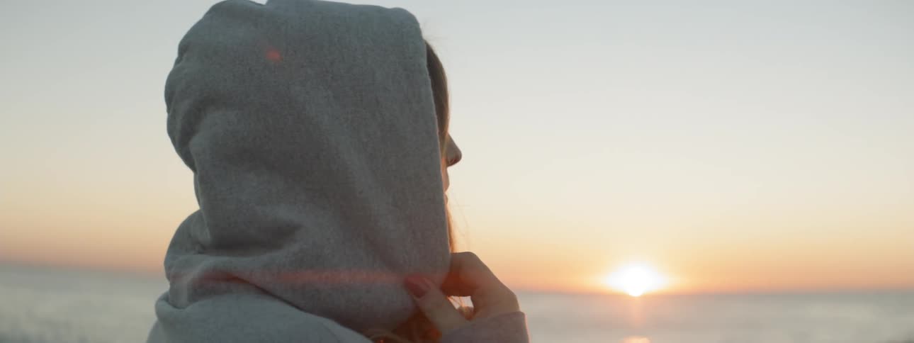 A woman places the hood of her shirt on while staring out at the water