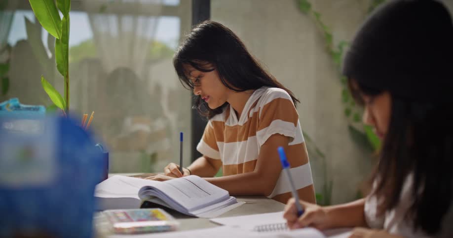 Two young people are sitting at a table writing in books and notebooks as they study together