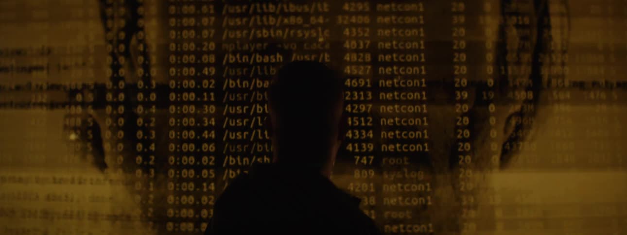 A man stands in front of a flickering screen displaying the image of a bearded man