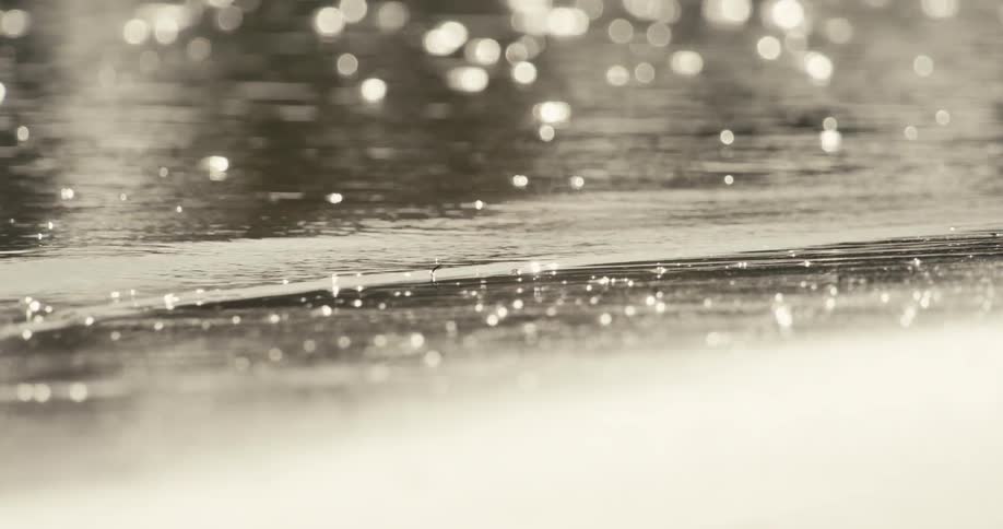 Sunlight sparkles and reflects off the gently rippling surface of shallow water creating a shimmering abstract effect with a receding wave