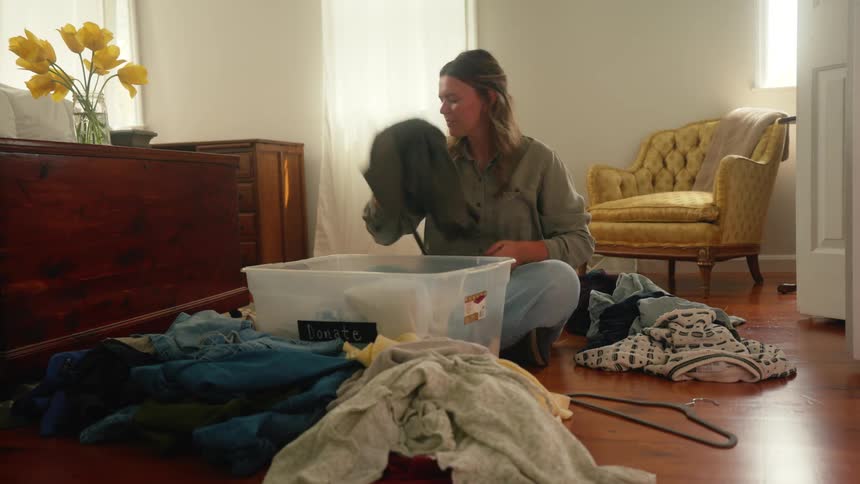 Woman sitting on the floor in her living room during the day surrounded by a pile of clean laundry as she folds the clothes and puts them in to the plastic bin in front of her.