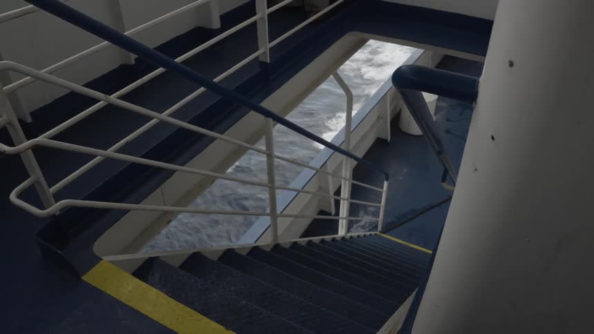 Looking down a set of metal stairs on a moving ship the churning ocean wake is visible through the railings and an opening to the side