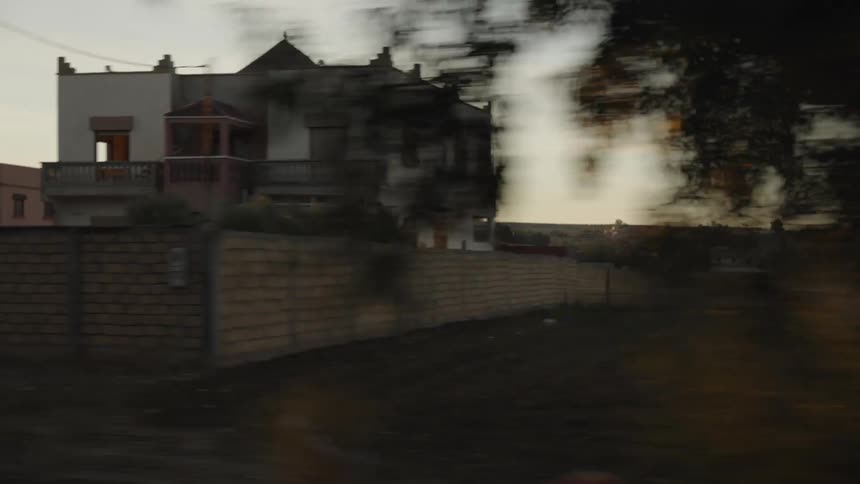 A blurry sideview from a moving vehicle shows a modest building in a rural area at sunset or sunrise