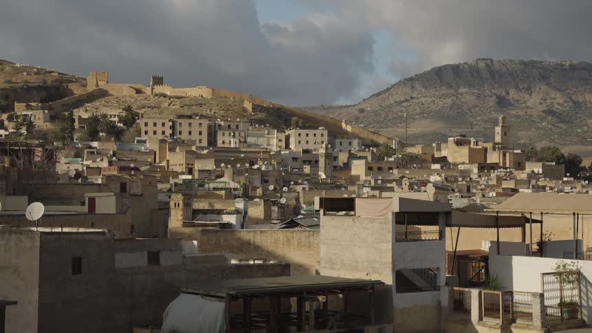 A slow pan reveals a city with densely packed buildings sprawling across a hillside under a partly cloudy sky with mountains visible in the background