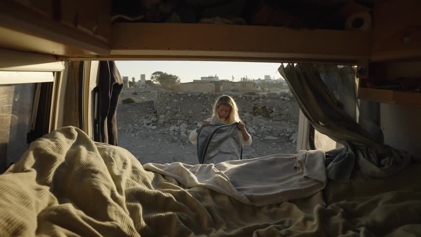 A woman unpacks a surfboard beside a van in a barren rocky area seen from inside the van