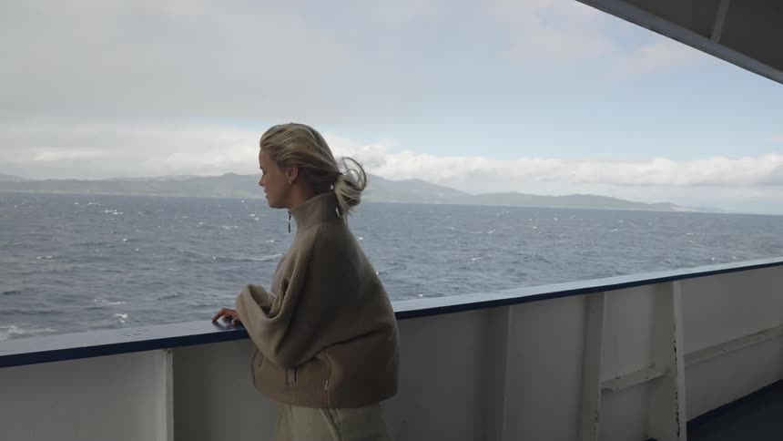 A woman stands on the deck of a boat as it moves through the ocean with the wind blowing her clothes and hair on a cloudy day