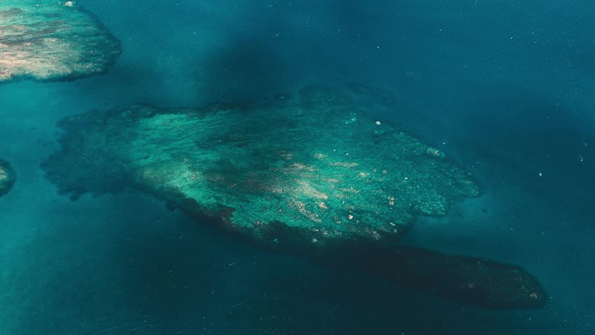 An aerial view captures a shallow reef or underwater landmass surrounded by deep blue ocean waters