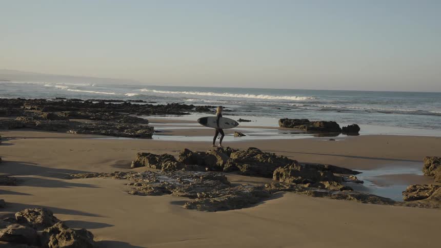 A lady walks towards the ocean with a surfboard at sunset