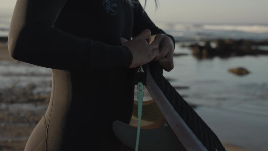 A woman in a wetsuit stands on a beach attaching a leash to her surfboard near the ocean