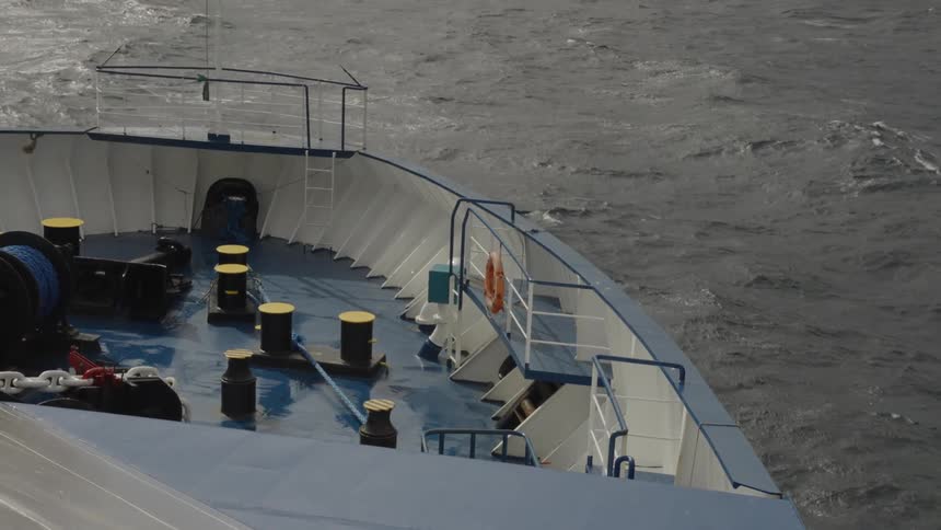 The front deck of a large ship is shown cutting through open water on an overcast day