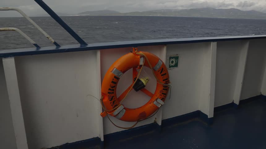 An orange lifebuoy is secured to the side of a moving ship with the choppy ocean and distant hazy land visible beyond the railing under a cloudy sky
