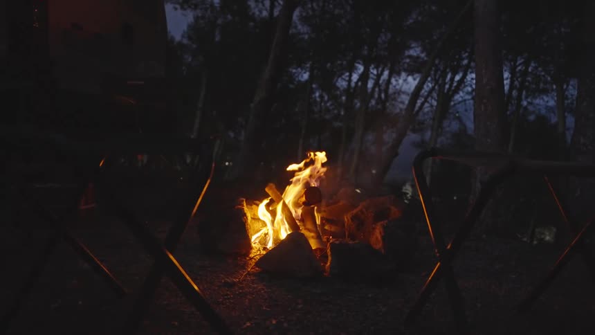 A campfire burns brightly at night in a forested campsite surrounded by empty folding chairs