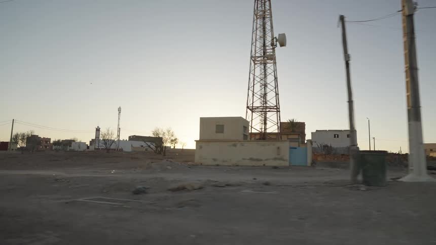 Captured from a moving vehicle the shot shows a quiet street at dawn in a developing neighborhood with the sun rising behind low buildings and a telecom tower