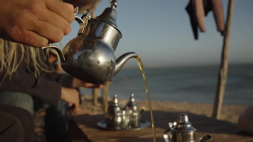 A person pours tea from a traditional metal teapot into a glass at a beachside setting