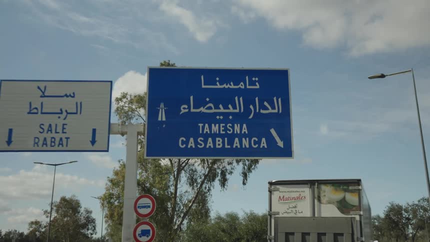 From a moving vehicle the view zooms in on large highway signs in arabic and english indicating directions to tamesna and casablanca as the signs pass overhead