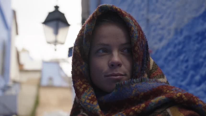 A woman wrapped in a colorful patterned shawl walks through a narrow blue painted alleyway glancing contemplatively to the side