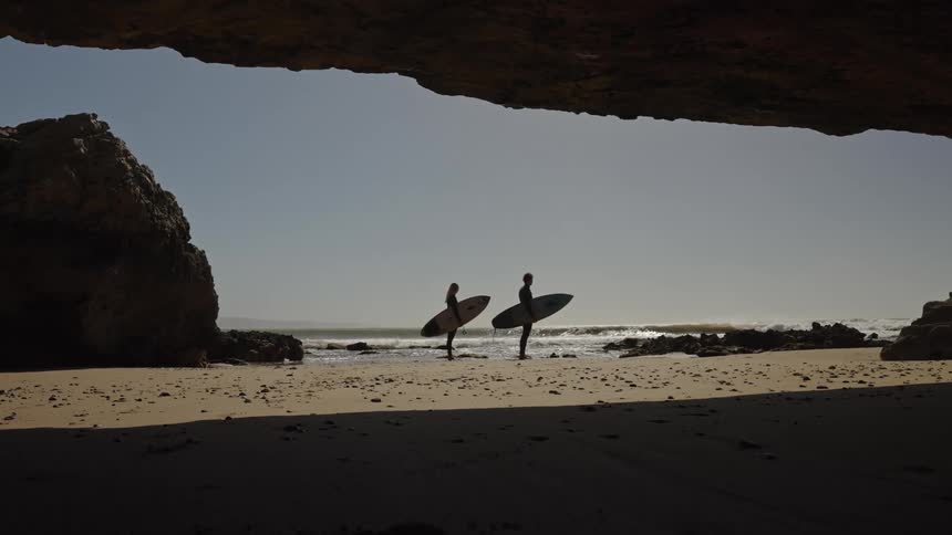Looking out from cave to pair of surfers holding surfboards on shoreline at the beach