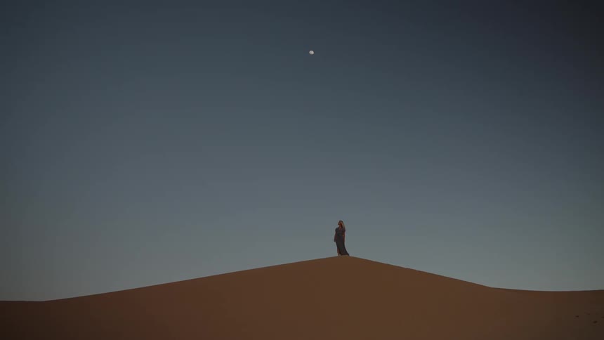 Woman wearing a long dress stands on a sand dune beneath the moon